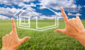 Bilda av två händer som ramar in ett hus som är placerat på grönt gräs med blå himmel.
